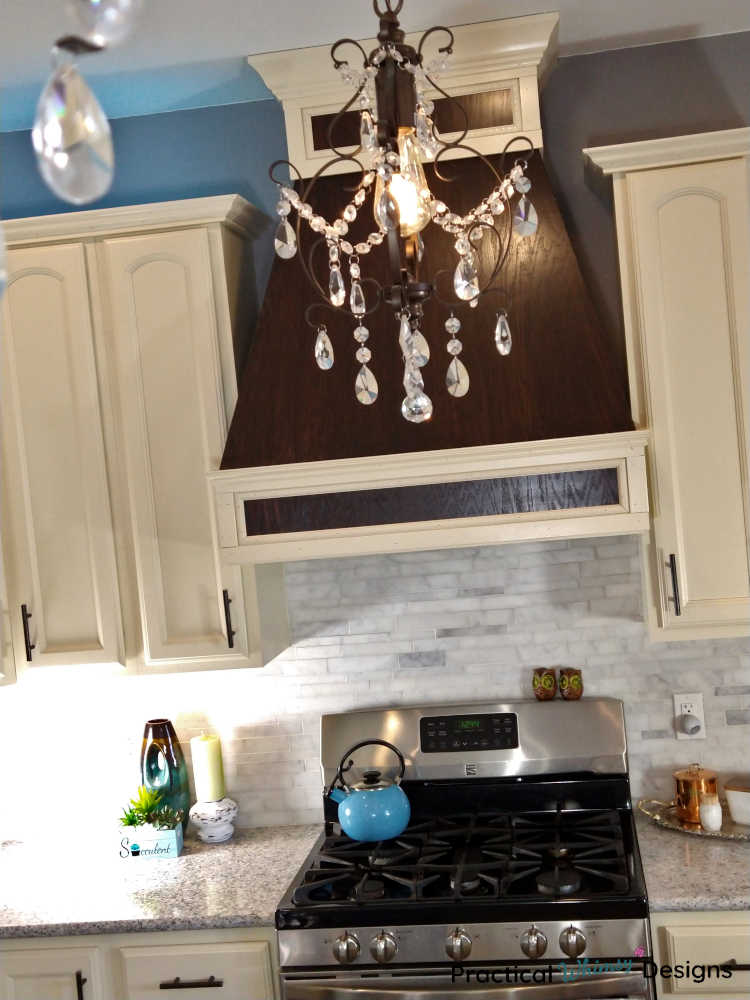 Glass chandelier hanging in front of custom wooden stove vent in remodeled kitchen