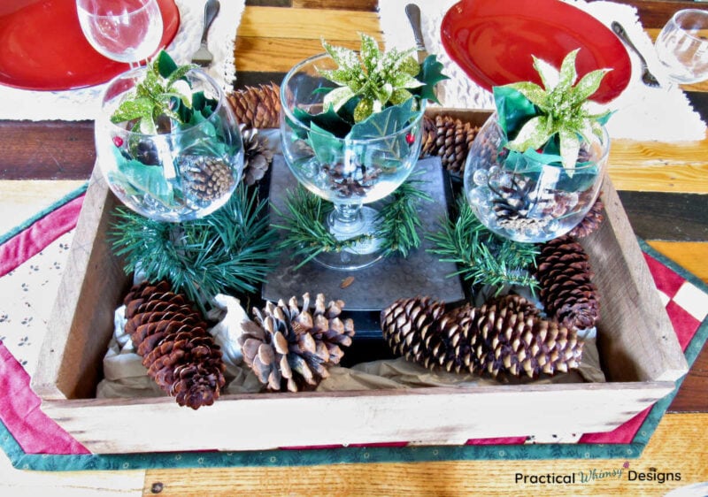 Wooden crate filled with pinecones, flowers, and clear glasses on table