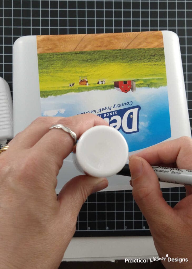 Hands drawing a circle on an empty ice cream box using a permanent marker and top of a small paint bottle.