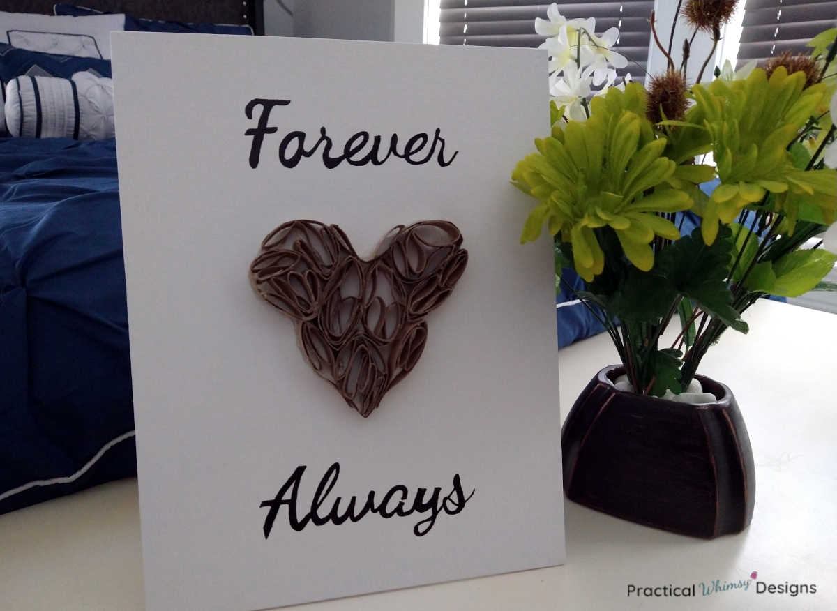 Heart toilet paper roll art on dresser next to green flowers in vase