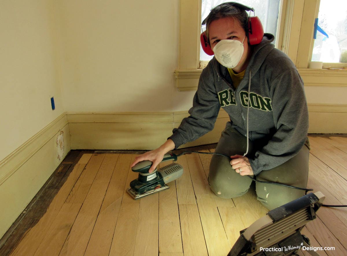 Lady in dust mask sanding the wooden floor with a hand sander.