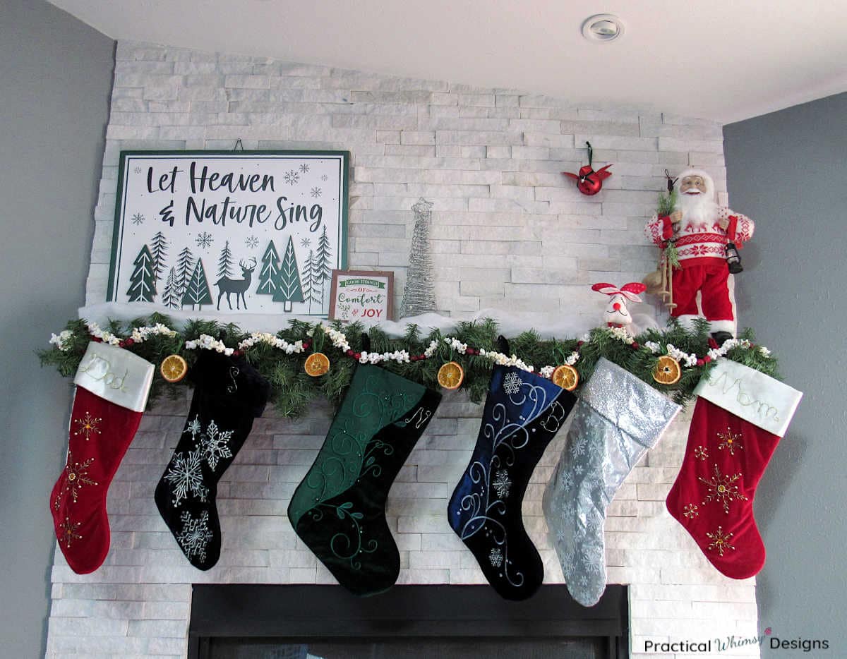 Old fashioned Christmas mantel decorated with signs, stockings, santa, and greenery.