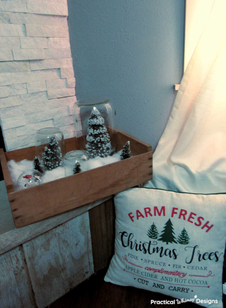 Pine tree snow globes in crate on hearth next to Christmas tree pillow.