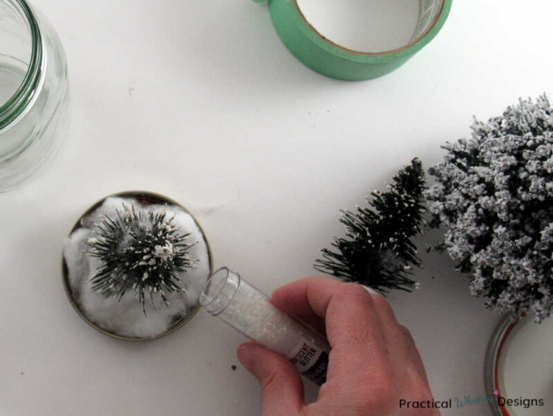 Dumping glitter on fake snow around a bottle brush tree in a jar lid.