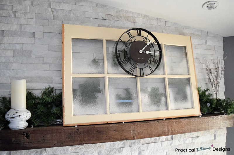 Spray snow window with brown clock and evergreens on mantel.