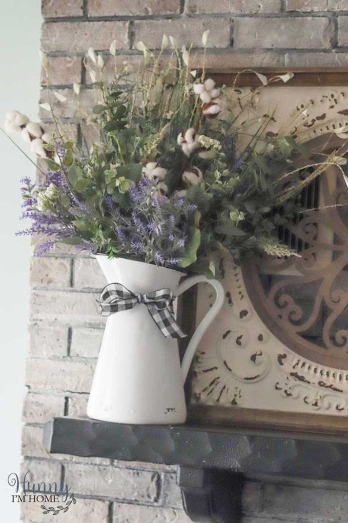 White pitcher with checkered ribbon filled with flowers sitting on a mantel.