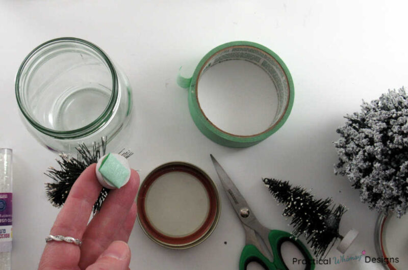 Supplies for pine tree snow globe and hand holding tree with tape stuck to the bottom.